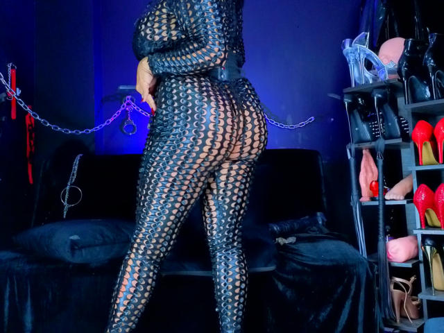 ScarletteStones in a black, net-like bodysuit, showing her butt and legs, with chains in the background, and a shelf with sex toys on the right.