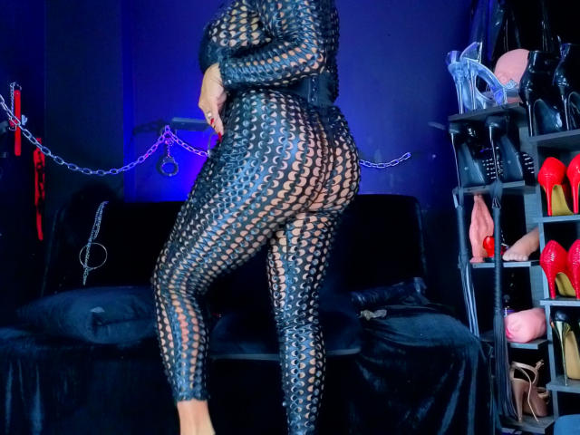 ScarletteStones in a black, wavy-patterned, see-through bodysuit, showing her ass. She's standing in a dark room with blue lighting, black velvet couch, and a shelf with red heels.