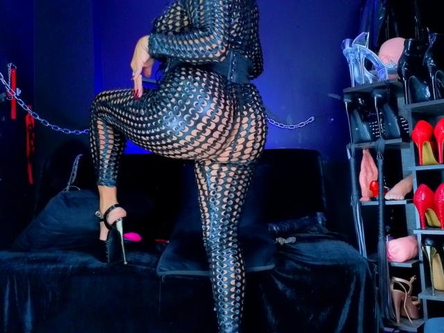 ScarletteStones in a black, fishnet bodysuit with her ass and pussy visible, wearing high heels, standing with one leg up, in a dark, blue-lit room.