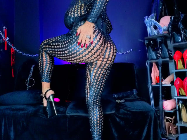 ScarletteStones in black fishnet bodysuit with red nails, standing with one leg up, showing her big ass, in high heels, in a dark, blue-lit room with BDSM gear.