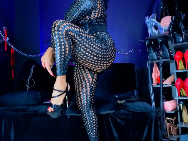 ScarletteStones in black fishnet lingerie with large circular holes, black high heels, raising one leg, showing her ass, in a blue-lit room with BDSM gear and shoes.