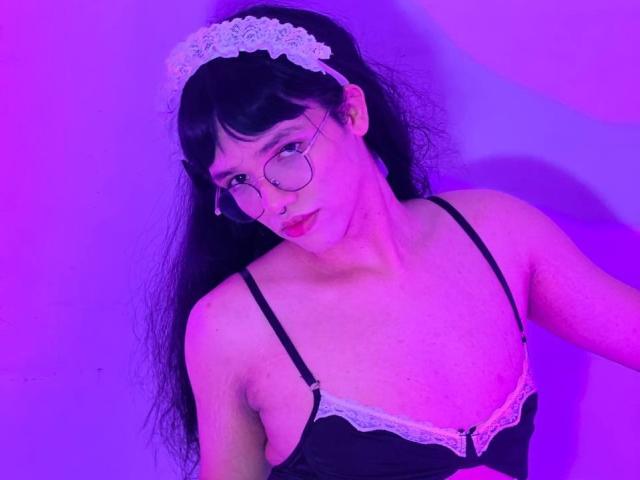 LuaGrey with long black hair, lace headband, round glasses, and black lace bra, purple-tinged lighting, looking up with a slight smile, showing small tits and a cute face.