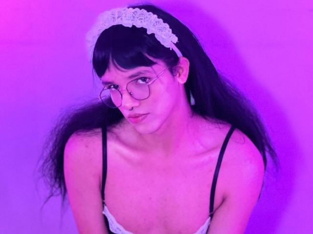LuaGrey with long black hair, lace headband, glasses, and white lingerie, showing small tits and a cute face, under purple lighting. She looks down with a slight pout.