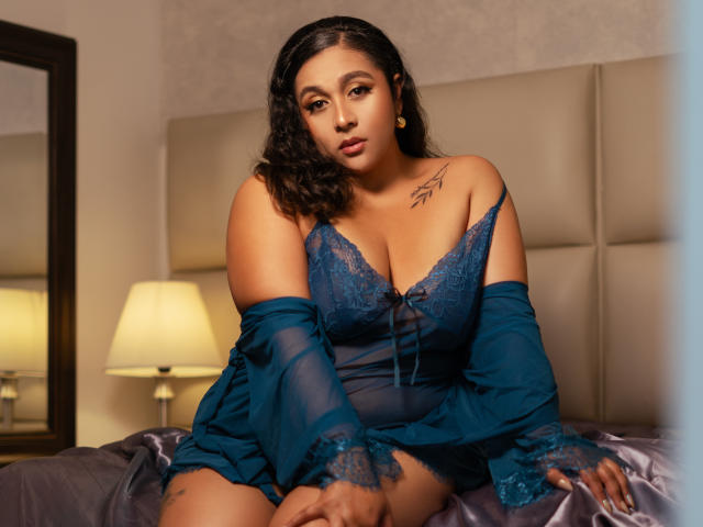 ViickyLee with curvy, tan-skinned body, large tits in blue lace lingerie, off-shoulder top, tattoo on left shoulder, sitting on bed, looking at camera, warm bedroom light.