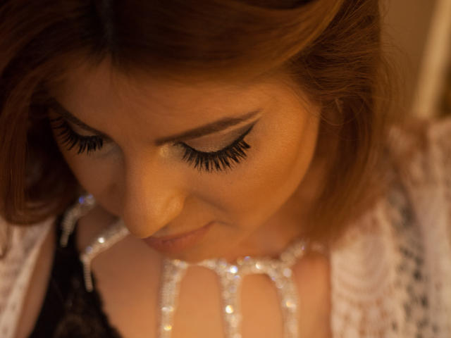 Le visage de Sexydollhotx, ses cheveux bruns et ses yeux fermés avec de longs cils noirs, regardant vers le bas des bijoux blancs étincelants sur ses mains, portant un haut en dentelle, un éclairage doux et chaleureux..