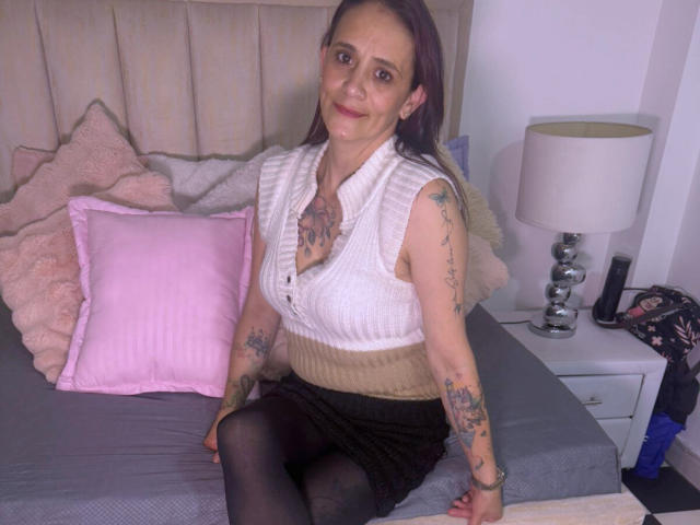 AmandaStoneX with tattoos, white sleeveless top, brown skirt, black tights, sitting on bed, pink pillow, beige headboard, gray bed, white lamp, smiling, medium tits, long brown hair.