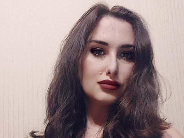Ambifadora has long, wavy brown hair, full red lips, and dark, subtle eye makeup. She's looking straight at the camera with a slight, confident smile.