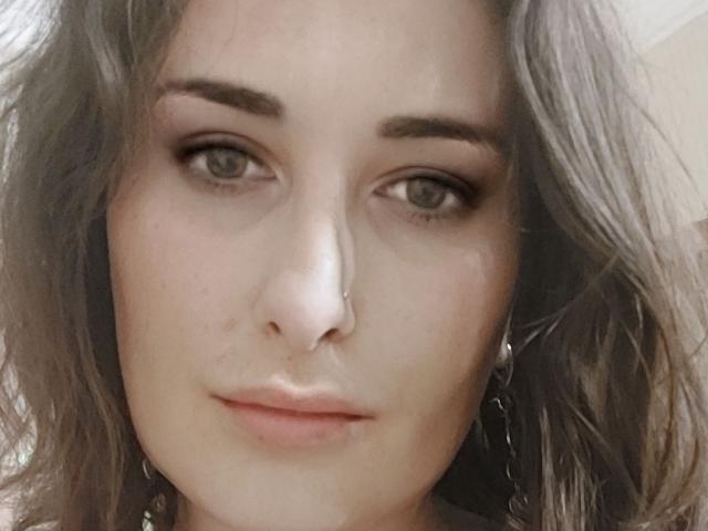 Ambifadora has light skin, wavy brown hair, green eyes, and a nose ring. She's looking directly at the camera with a slight, closed-lip smile. Her eyebrows are dark and thick.