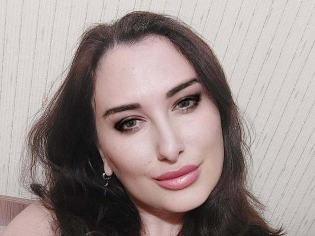 Ambifadora has dark brown wavy hair, green eyes, thick eyebrows, and pink lipstick. She's smiling with a slight nose piercing and small hoop earrings, looking cute and sexy.