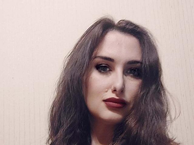 Ambifadora has wavy brown hair, dark red lipstick, and subtle makeup with dark eyeliner, staring directly with a slight, confident smile. Her skin is fair.
