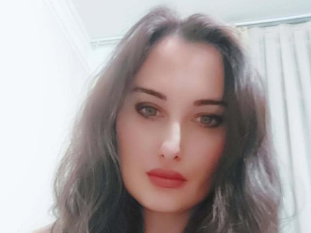Ambifadora has wavy dark brown hair, green eyes, and red lipstick, with a slightly blurred, soft-focused selfie showing her face and upper chest.