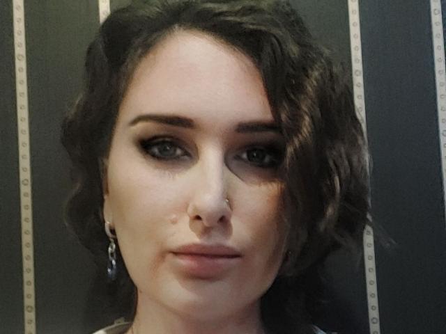 Ambifadora has wavy brown hair, a nose ring, and dark eyeliner. Her small tits are slightly visible under her shirt. She wears earrings and has a serious expression.