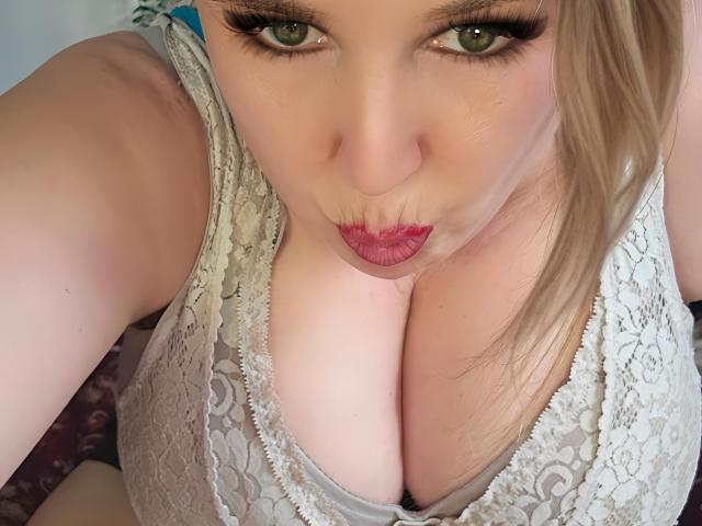 TitanicTits with puffy pink lips, green eyes, and blonde hair, looking up at the camera, wearing a lace top that highlights her big, round, pale tits.