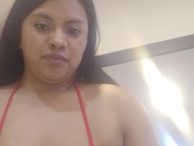 DahianaColeman with medium brown skin, long black hair, and red halter top, showing her bare shoulders and subtle cleavage, giving a slightly pouty expression.