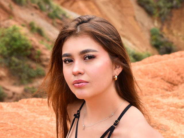 VioletaConnors with brown hair, nose ring, and gold earrings, wearing a black strappy top, has a serious expression, and a slight pout, with a tan, rocky background.