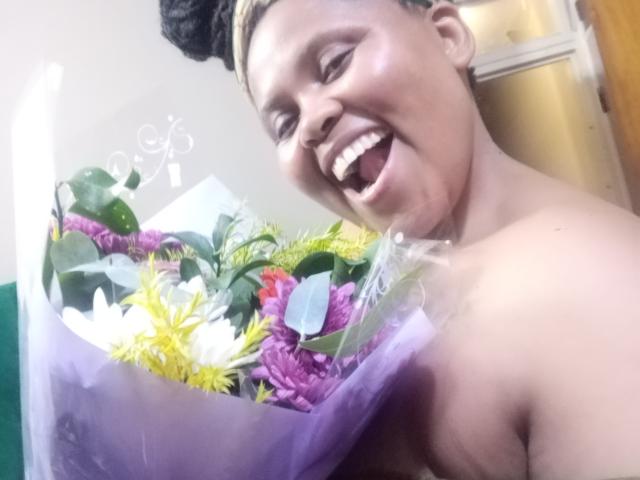 Thicknesdx with dark skin and braided hair, smiling with tongue out, holding a colorful bouquet of flowers in translucent paper. Topless, showing small to medium tits.