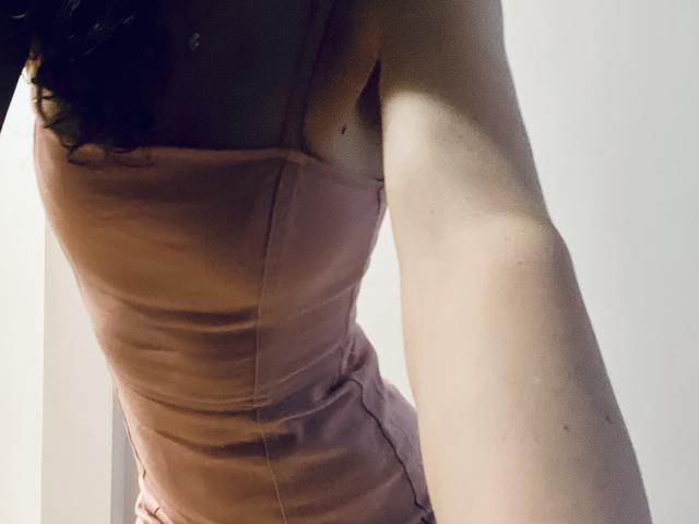 De roze jurk van SweetClaraa laat haar kleine borsten en blote arm zien. Haar krullende haar is gedeeltelijk zichtbaar. Ze heeft een lichte huidskleur. De afbeelding is een foto..