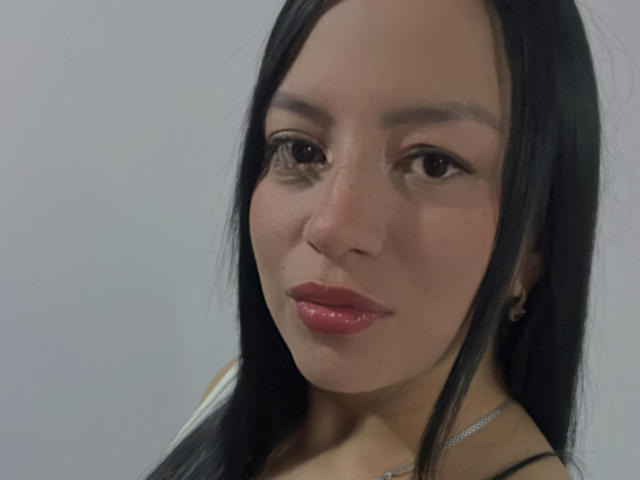 MiiaG has long black hair, brown eyes, and shiny pink lips. She's wearing a silver necklace, and her face is centered in the shot. Her skin is smooth and light.