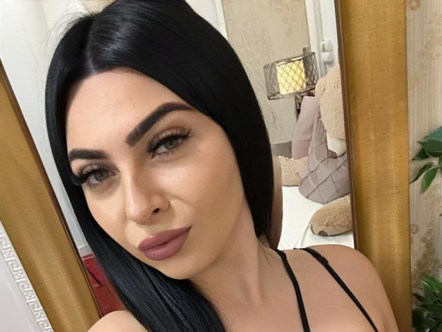 RileySinclair has black hair, green eyes, and full pink lips. She's wearing a black spaghetti strap top, looking at the camera with a slight smile.