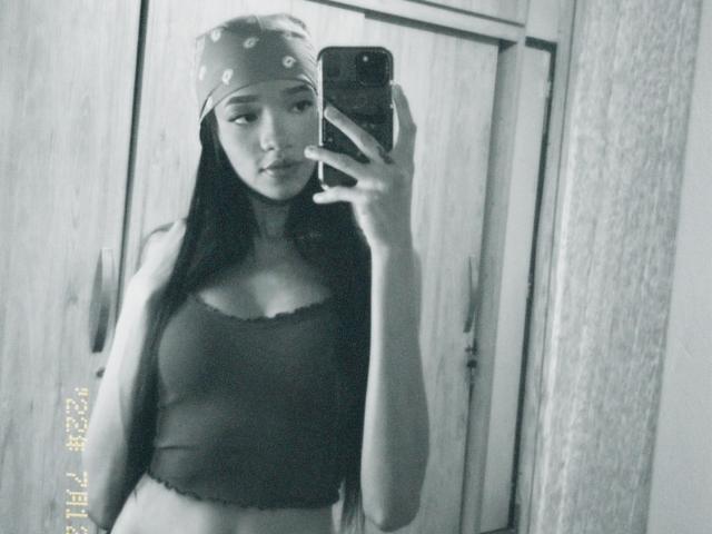 Black-and-white selfie of IrinaDrew with long dark hair, wearing a polka-dot headscarf, tank top showing small tits, holding phone, standing in front of wooden wardrobe.