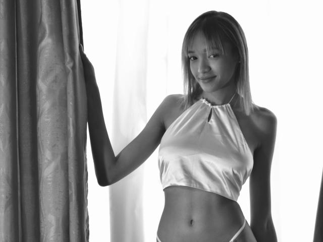 Foto em preto e branco de IrinaDrew, com seios pequenos, vestindo um top de alça prateado brilhante e uma tanga branca, em pé perto de uma cortina, sorrindo discretamente..