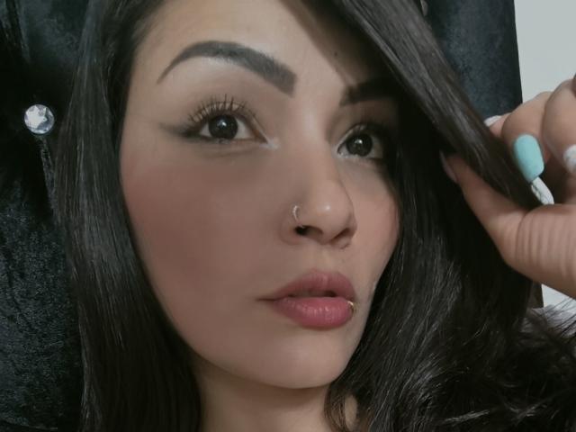 AliceGiorgia has dark brown hair, a nose ring, and a lip piercing. She's touching her hair with a blue-painted nail, looking slightly to the right.