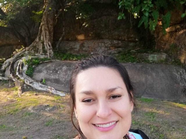 LadySelene with medium brown skin, dark brown eyes, and a small smile, wearing a black top, has her dark hair pulled back, standing in a sunlit, rocky outdoor area.