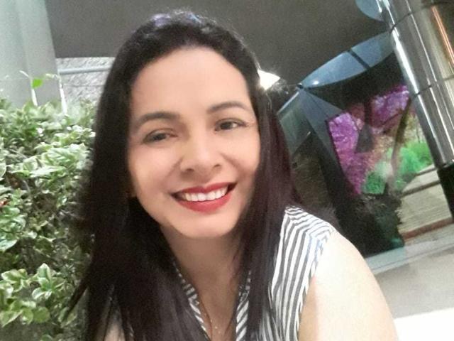 PamelaSmiths with long black hair, smiling with red lipstick, wearing a striped sleeveless top, showing white teeth, and standing next to a green plant.