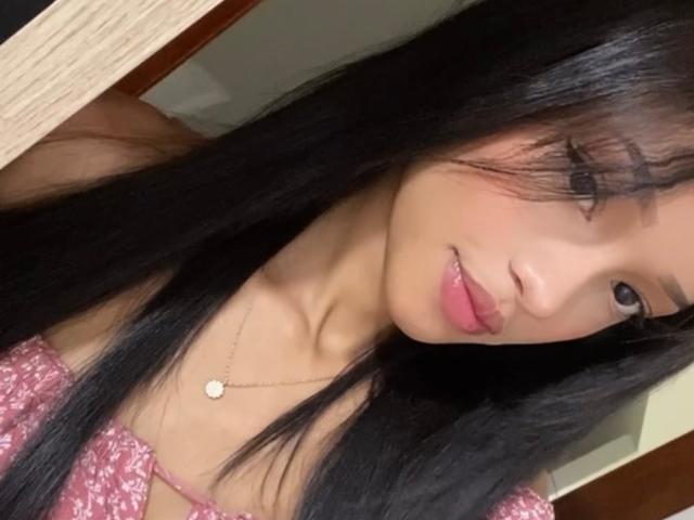 IrinaDrew with long black hair, pink sparkly top, and pink lipstick, looking at camera with a slight smile, wearing a small gold necklace. Her tits are small and cute.