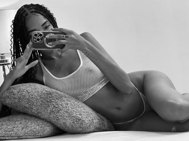 IrinaDrew with curly hair, wearing a mesh sports bra and tiny panties, lying on pillows, taking a mirror selfie with her phone, showing her tits and pussy. Black-and-white photo.