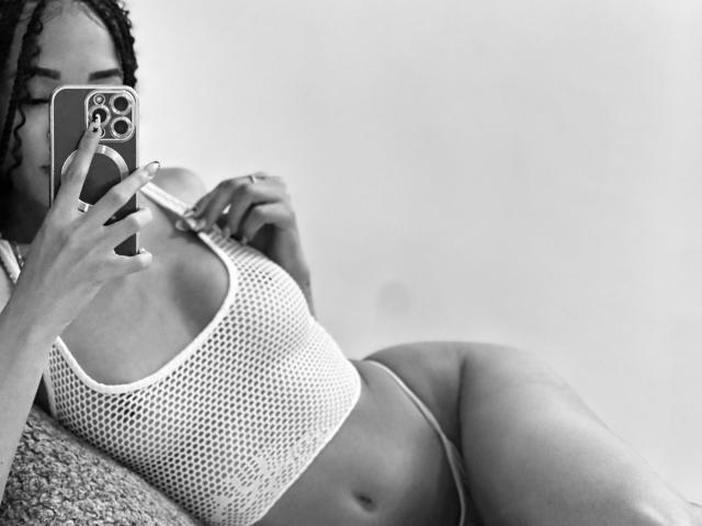 Black-and-white selfie of IrinaDrew with braided hair, holding phone, wearing mesh sports bra, showing small tits, and thong, lying down, showing flat stomach and ass.