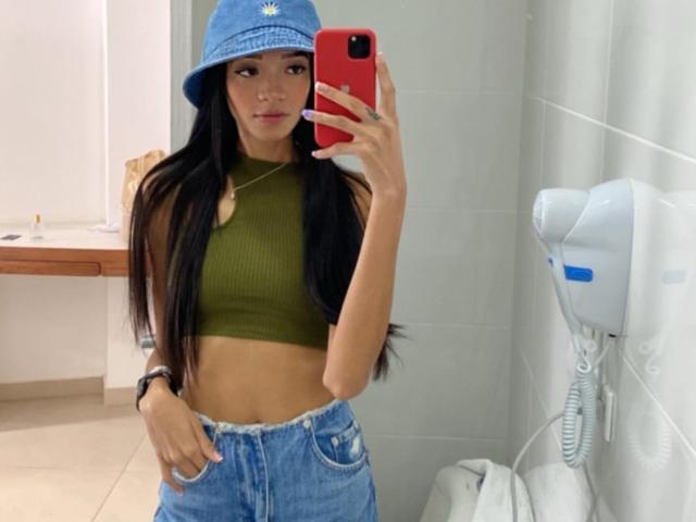 IrinaDrew, avec de longs cheveux noirs, portant un chapeau de pêche bleu, un crop top vert et un jean bleu, tenant un téléphone rouge, montrant un ventre plat et de petites seins..