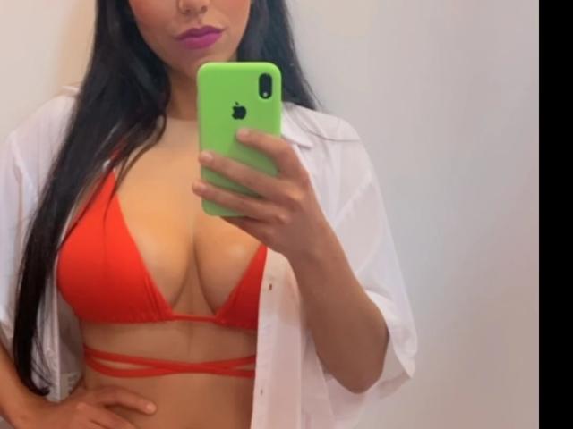 Kim69x che si fa un selfie, mostrando un seno prosperoso con un top da bikini rosso con spalline, indossando una camicia bianca aperta, tenendo un iPhone verde, con lunghi capelli neri e labbra viola..