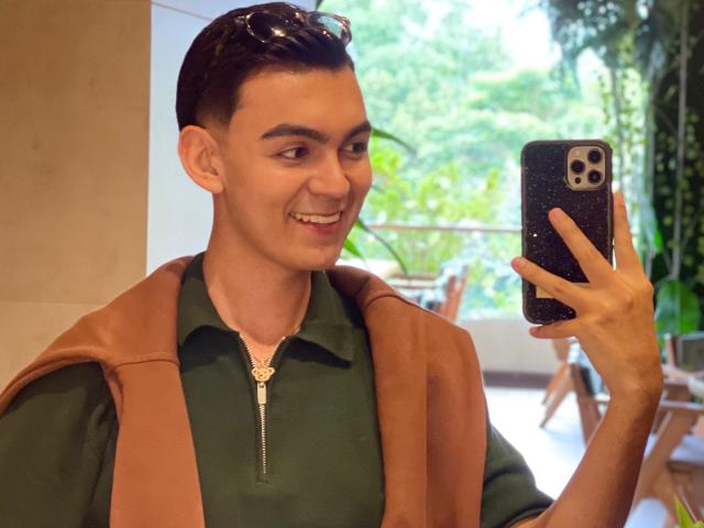 DeusBeauty, smiling, holding a phone, wearing a green polo with a brown vest, short dark hair, slightly tanned, looking at the phone, standing indoors, taking a selfie.