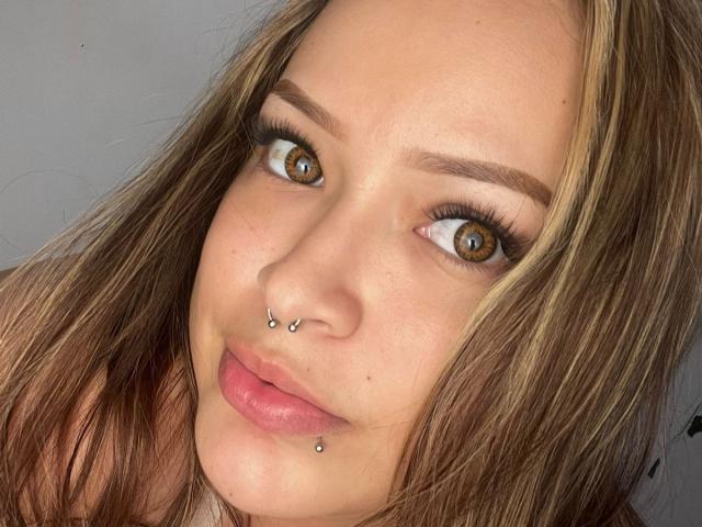 AllisonJustingb's face with brown eyes, blonde-brown hair, nose piercings, and lower lip piercing, looking at camera with pinkish lips, slight smile.