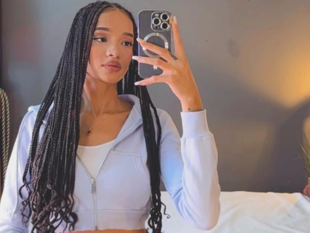 IrinaDrew with long black braids, white zip-up hoodie, and white top, holding a phone, taking a mirror selfie, showing her cute face and small tits.