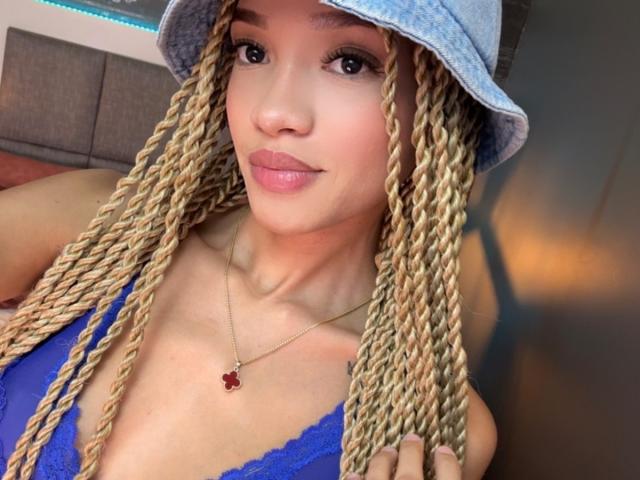 IrinaDrew with blonde braids, wearing a blue bucket hat and blue lace top, showing her tits with a red flower necklace, smiling at the camera.