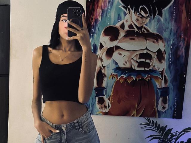 IrinaDrew with black crop top, jeans, and black beanie, mirror selfie showing muscular anime poster of Super Saiyan, hand in pocket, standing in dimly lit room.