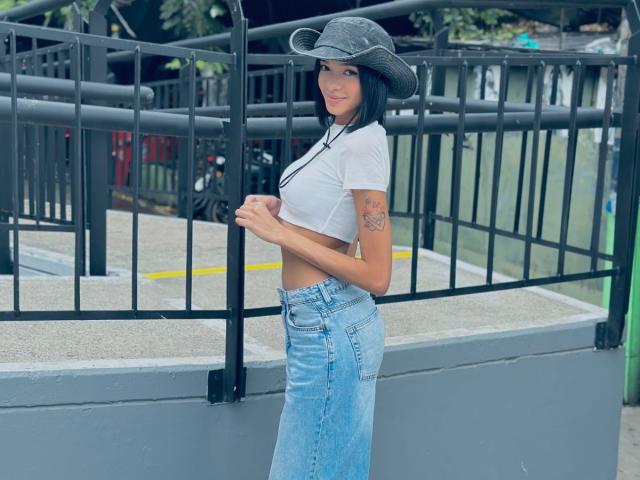IrinaDrew stands with a gray cowboy hat, white crop top, and blue high-waisted jeans, showing her tattooed arm and small tits, smiling at the camera.
