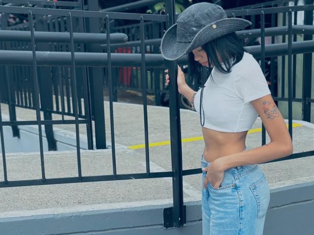 IrinaDrew with black hat, white crop top, blue jeans, tattoo on right arm, touching fence, looking down, hands in pockets, showing her small tits and flat ass.