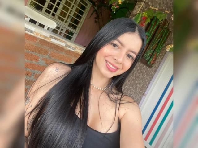 IsabellaJames has long black hair, a pearl necklace, and is smiling with pink lipstick, showing small tits and a tattoo on her right arm, in a selfie.