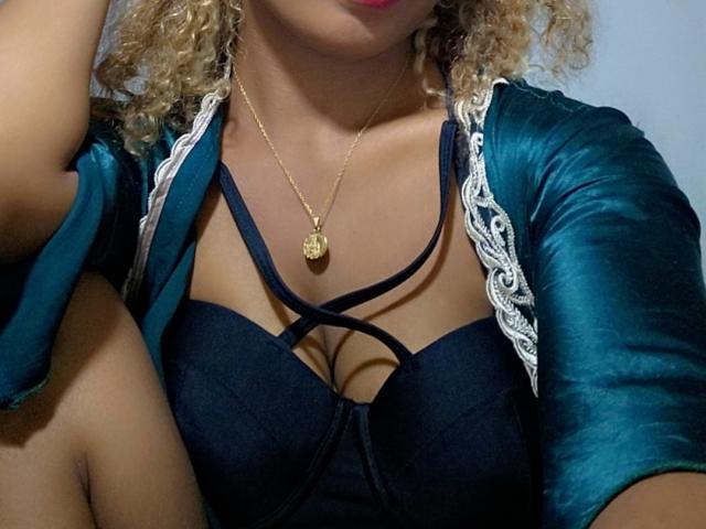 TiakoI, con cabello rubio rizado, vestida con una bata de satén color turquesa, un sujetador negro con tirantes, y un collar dorado, mostrando sus pechos y labios rojos, sentada con una rodilla levantada..