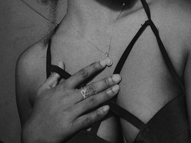 La mano de TiakoI, con un anillo en el dedo, toca sus pechos desnudos, vistiendo un sujetador negro con tirantes, un delicado collar con un pequeño colgante circular, en una fotografía en blanco y negro..