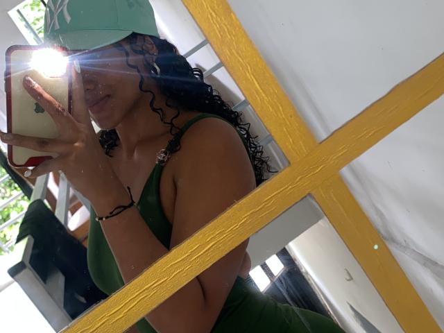 NatalyBlonder with curly black hair, green top, and green pants, taking a mirror selfie with a sun flare, wearing a green cap, showing her tits in the reflection.
