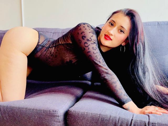 NancyRouse is on a gray couch, wearing a black, sheer leopard-print bodysuit, showing her ass, with long black hair, red lipstick, and hoop earrings, looking at the camera.