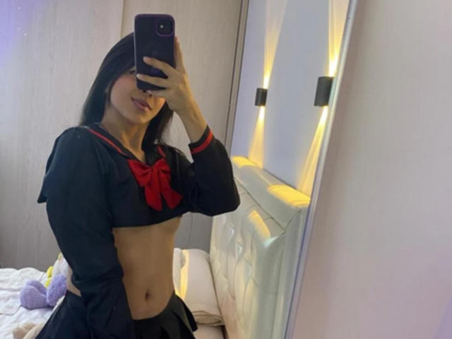 MagiicBebes vêtu d'une tenue de marin noire avec un nœud rouge, montrant un ventre plat et des cheveux noirs, prenant un selfie devant un miroir avec un téléphone, sans pantalon, révélant une petite vulve..