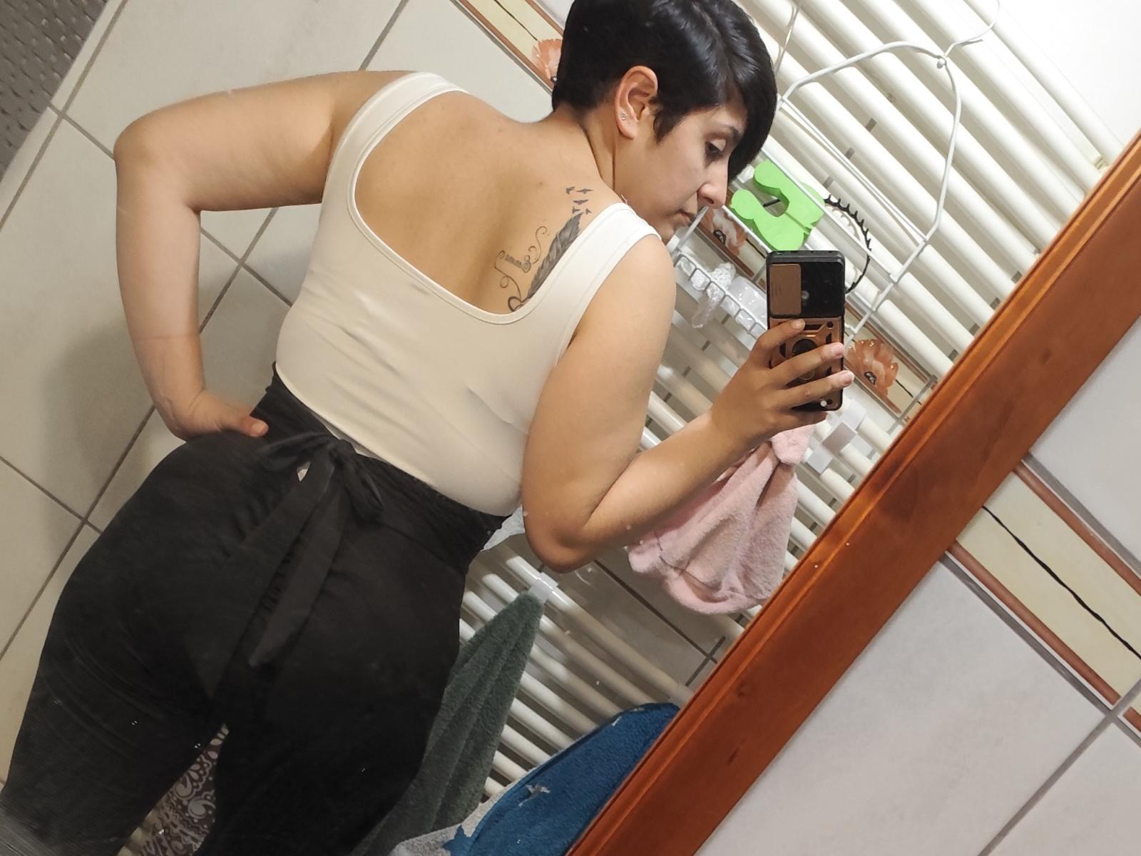 MarieChris69 with short black hair, white tank top, black pants, butt tattoo, taking mirror selfie, showing back, hand on hip, holding phone, towel on radiator, bathroom.