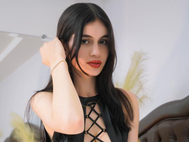 MarilynScott with long black hair, red lipstick, and a black lace-up dress, touching her hair with a gold bracelet on her wrist, showing her tits slightly.
