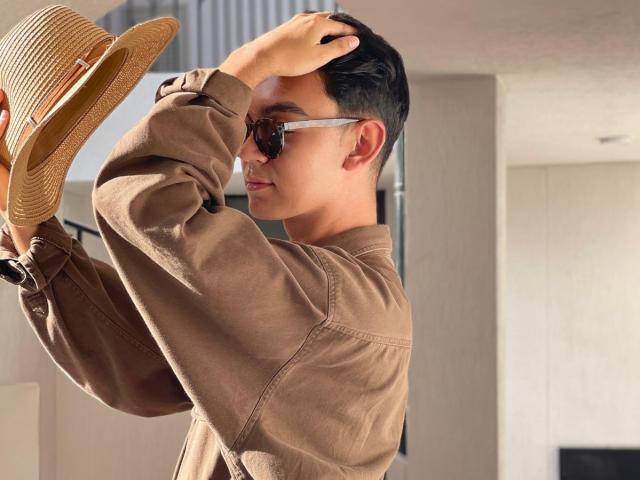 DeusBeauty in a brown jacket, holding a straw hat, wearing sunglasses, with short black hair, raising his hand to his head. Sunlight casts a warm, natural glow.