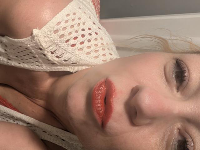 PowderHot liegt da, ihr Gesicht mit glänzenden roten Lippen, einem weißen Spitzenoberteil und blonden Haaren. Ihre Augen sind halb geschlossen, und ihre Haut ist blass mit einem leichten Glanz..